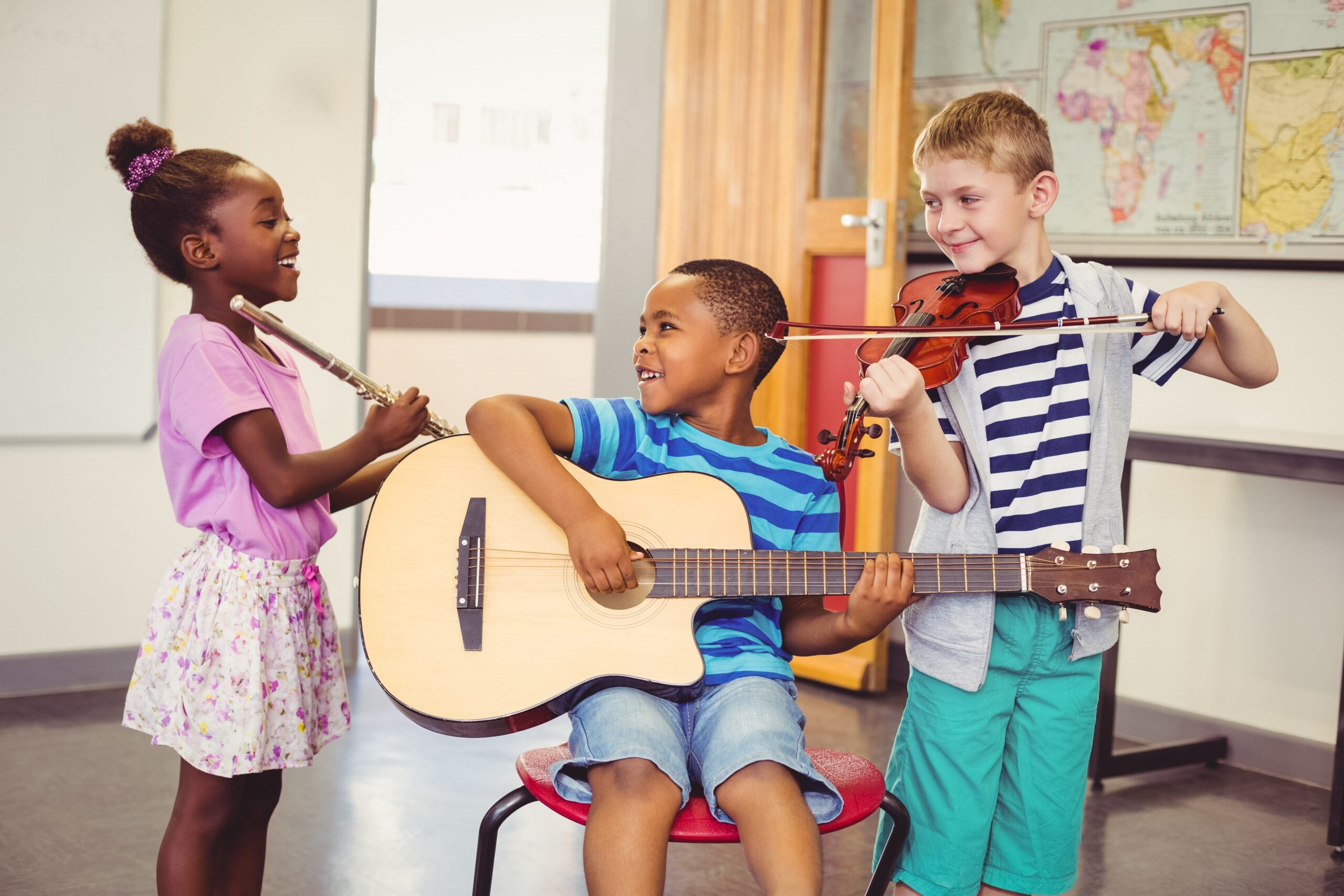 A trio of cheerful young children in a classroom play music together on a flute, guitar, and violin