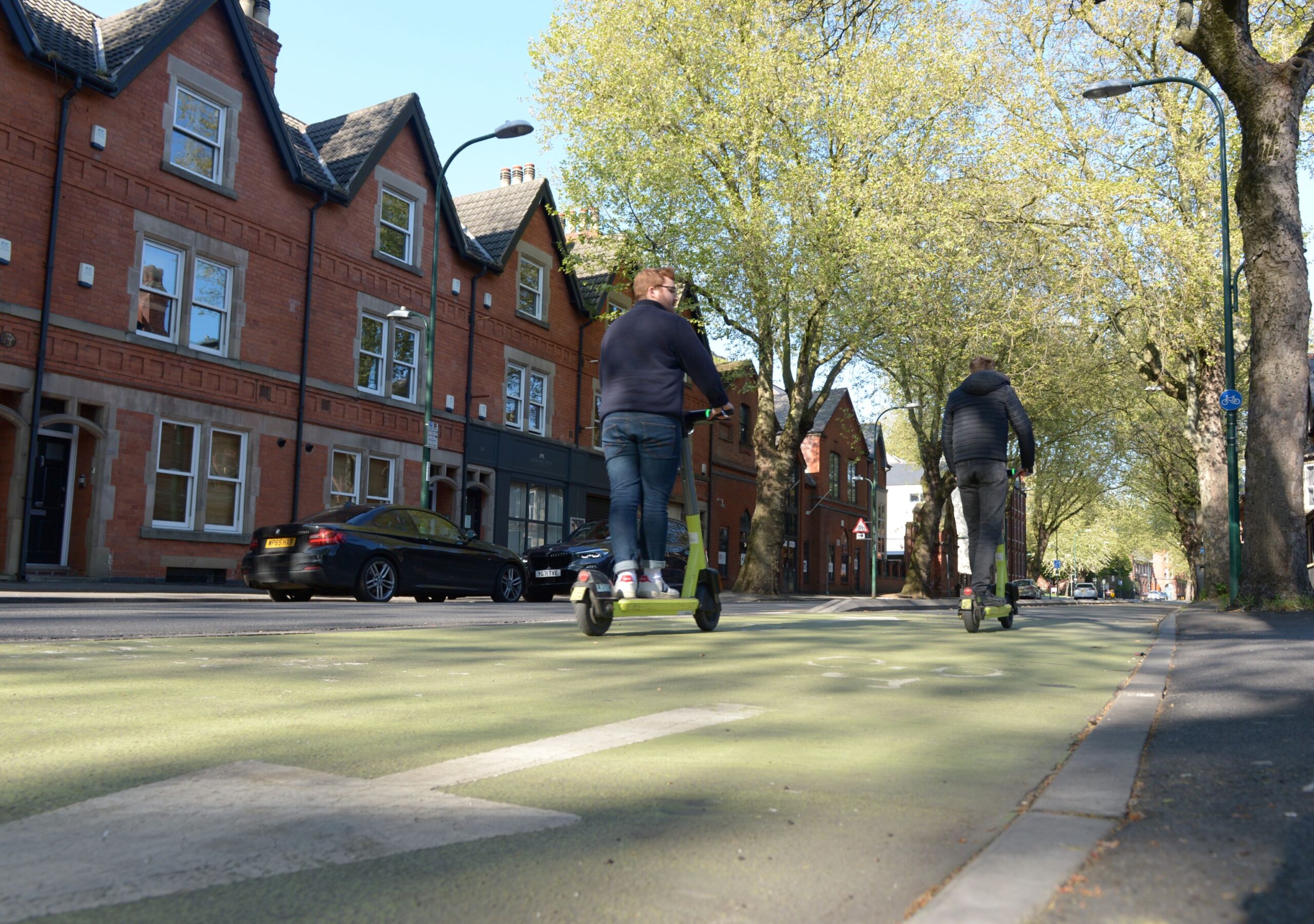 A view of two young men riding e-scooters along a bike path on a tree-lined residential street on a sunny day.