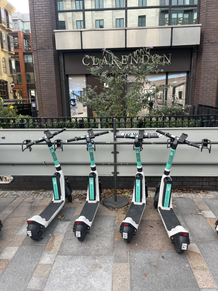 A row of 4 turquoise e-scooters parked in a row along an e-scooter parking bay on a paved public walkway. 